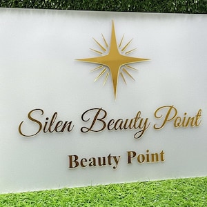 May include: A rectangular sign with the words "Silen Beauty Point" and "Beauty Point" in gold lettering. A gold starburst design is at the top. The sign is on a white background.