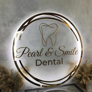 May include: A circular, backlit wall sign featuring a frosted glass surface with a gold-toned outline of a tooth at the top. The text "Pearl & Smile" is written in a gold cursive font, with the word "Dental" in a gold sans-serif font below.