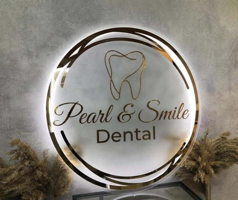 May include: A circular, backlit wall sign featuring a frosted glass surface with a gold-toned outline of a tooth at the top. The text "Pearl & Smile" is written in a gold cursive font, with the word "Dental" in a gold sans-serif font below.