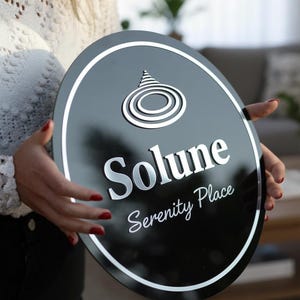 May include: A black oval sign with a white border and silver lettering. The sign reads "Solune Serenity Place" and features a stylized design at the top. The sign is held by a person.