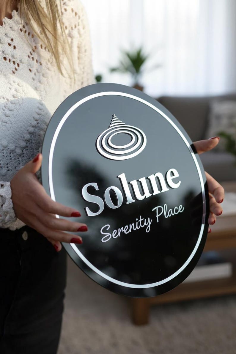 May include: A black oval sign with a white border and silver lettering. The sign reads "Solune Serenity Place" and features a stylized design at the top. The sign is held by a person.