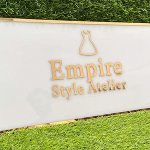 May include: A rectangular sign with a white background and gold trim. The sign features the words "Empire Style Atelier" in gold lettering, along with a gold dress silhouette. The sign is set against a backdrop of green grass.