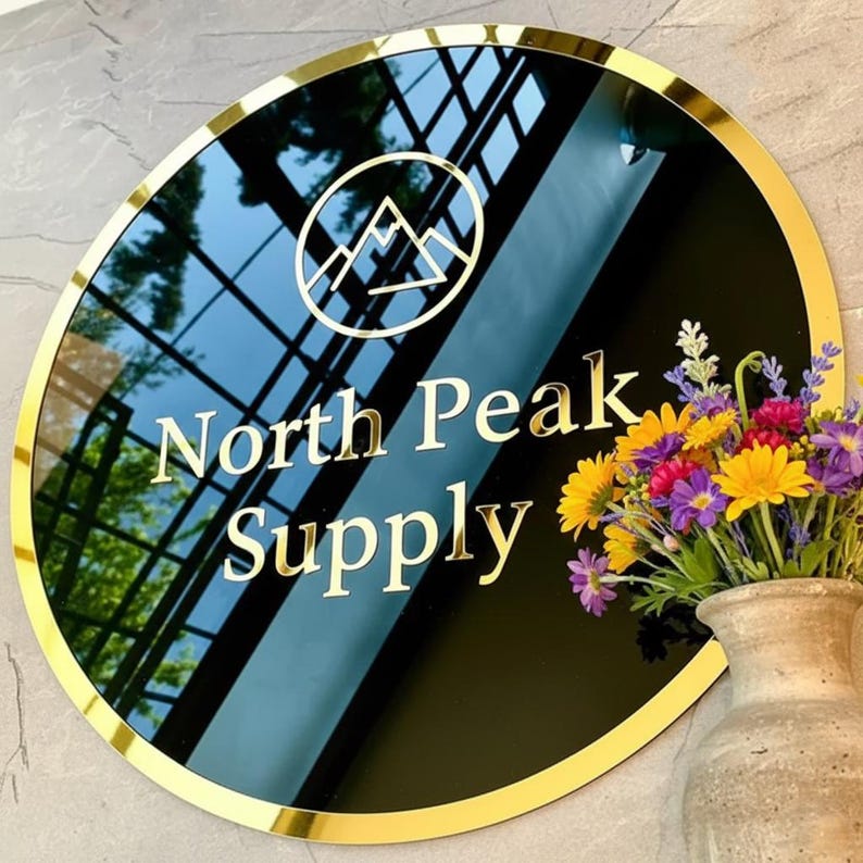 May include: A round black sign with gold trim and lettering that reads "North Peak Supply." The sign features a gold mountain logo and is displayed near a vase of colorful flowers.