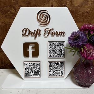 May include: A white, hexagonal sign with the words "Drift Form" in elegant script. It features a Facebook logo and three QR codes in a copper color. A copper spiral design is at the top. A vase of purple flowers sits to the right.
