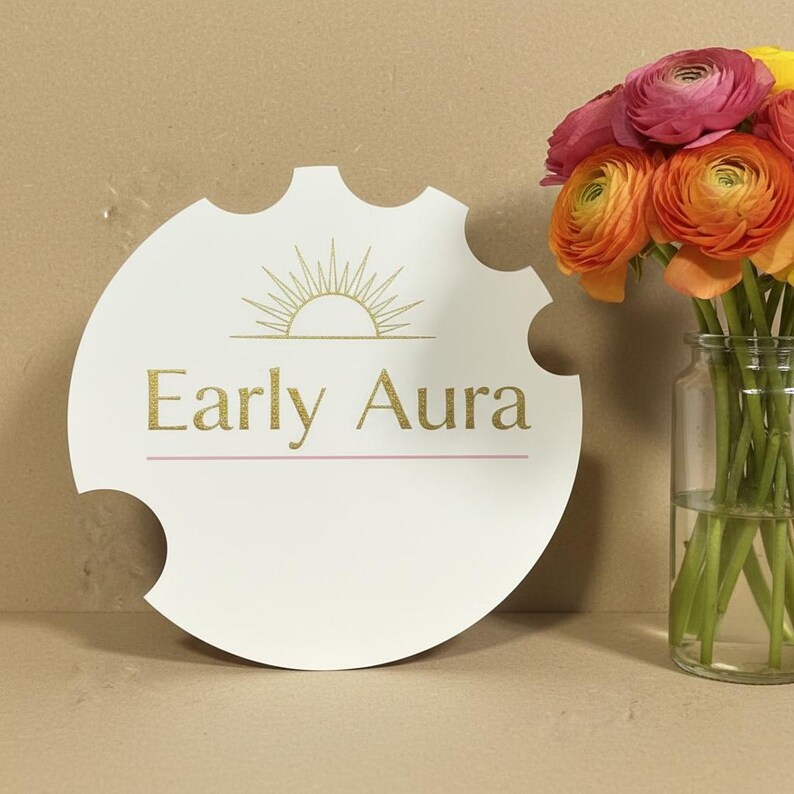 May include: A white, circular sign with a unique notched edge, featuring the text "Early Aura" in gold glitter lettering, and a gold sunburst design. A bouquet of orange, pink, and yellow flowers in a glass jar is to the right.