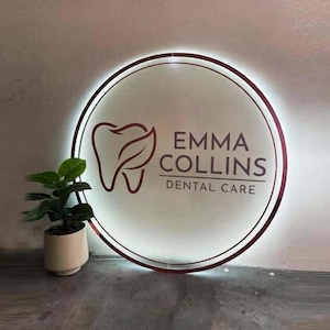May include: A circular, backlit wall sign featuring a dark maroon tooth icon with a leaf detail and the text EMMA COLLINS DENTAL CARE in dark maroon lettering. The sign has a glowing white border and is mounted on a wall.