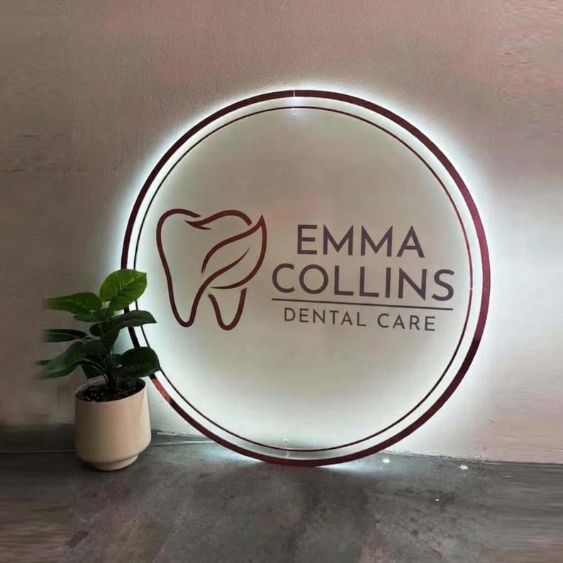May include: A circular, backlit wall sign featuring a dark maroon tooth icon with a leaf detail and the text EMMA COLLINS DENTAL CARE in dark maroon lettering. The sign has a glowing white border and is mounted on a wall.