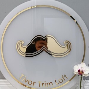 Pu&ograve; includere: Un cartello circolare bianco con un grafico a baffi dorato al centro e un bordo circolare dorato. Il testo Tivor Trim Loft &egrave; stampato in lettere dorate nella parte inferiore. Un'orchidea bianca &egrave; visibile in un vaso di vetro.