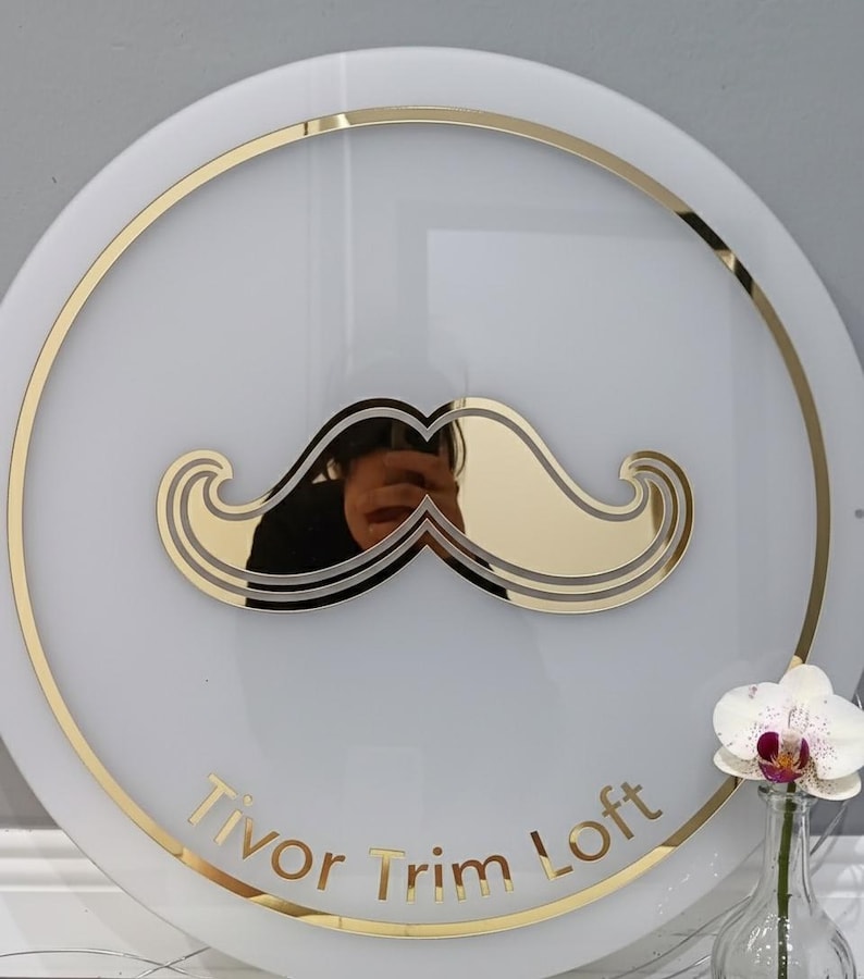 Pu&ograve; includere: Un cartello circolare bianco con un grafico a baffi dorato al centro e un bordo circolare dorato. Il testo Tivor Trim Loft &egrave; stampato in lettere dorate nella parte inferiore. Un'orchidea bianca &egrave; visibile in un vaso di vetro.