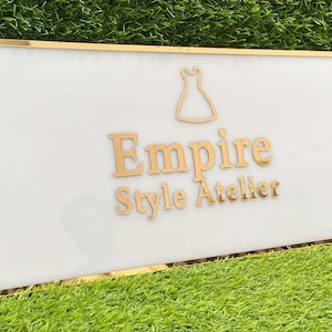 May include: A rectangular sign with a white background and gold trim. The sign features the words "Empire Style Atelier" in gold lettering, along with a gold dress silhouette. The sign is set against a backdrop of green grass.