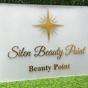 May include: A rectangular sign with the words "Silen Beauty Point" and "Beauty Point" in gold lettering. A gold starburst design is at the top. The sign is mounted on a white background.