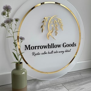 May include: A circular frosted acrylic sign with a gold metallic border. The sign features a gold willow branch logo and the black text "Morrowhllow Goods" followed by the phrase "Rustic calm built into every detail." in a script font.