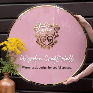 May include: A circular pink acrylic sign with a gold metallic border and a central gold crest featuring a flame icon. The text reads Wyrdon Craft Hall and Warm rustic design for soulful spaces in white and black lettering.