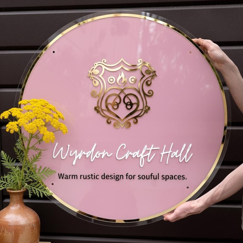 May include: A circular pink acrylic sign with a gold metallic border and a central gold crest featuring a flame icon. The text reads Wyrdon Craft Hall and Warm rustic design for soulful spaces in white and black lettering.