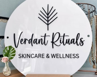 May include: A circular white acrylic sign featuring a black leaf icon, the text "Verdant Rituals" in a cursive font, and the text "SKINCARE & WELLNESS" in a sans-serif font. The sign is mounted on a rectangular wooden base.
