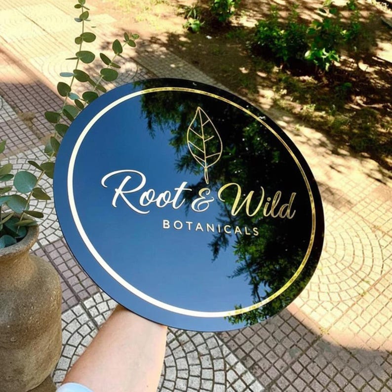 May include: A round, black sign with gold trim and lettering. The sign reads "Root & Wild BOTANICALS" in elegant script and features a gold leaf design. The sign is held up against a background of greenery and a paved surface.