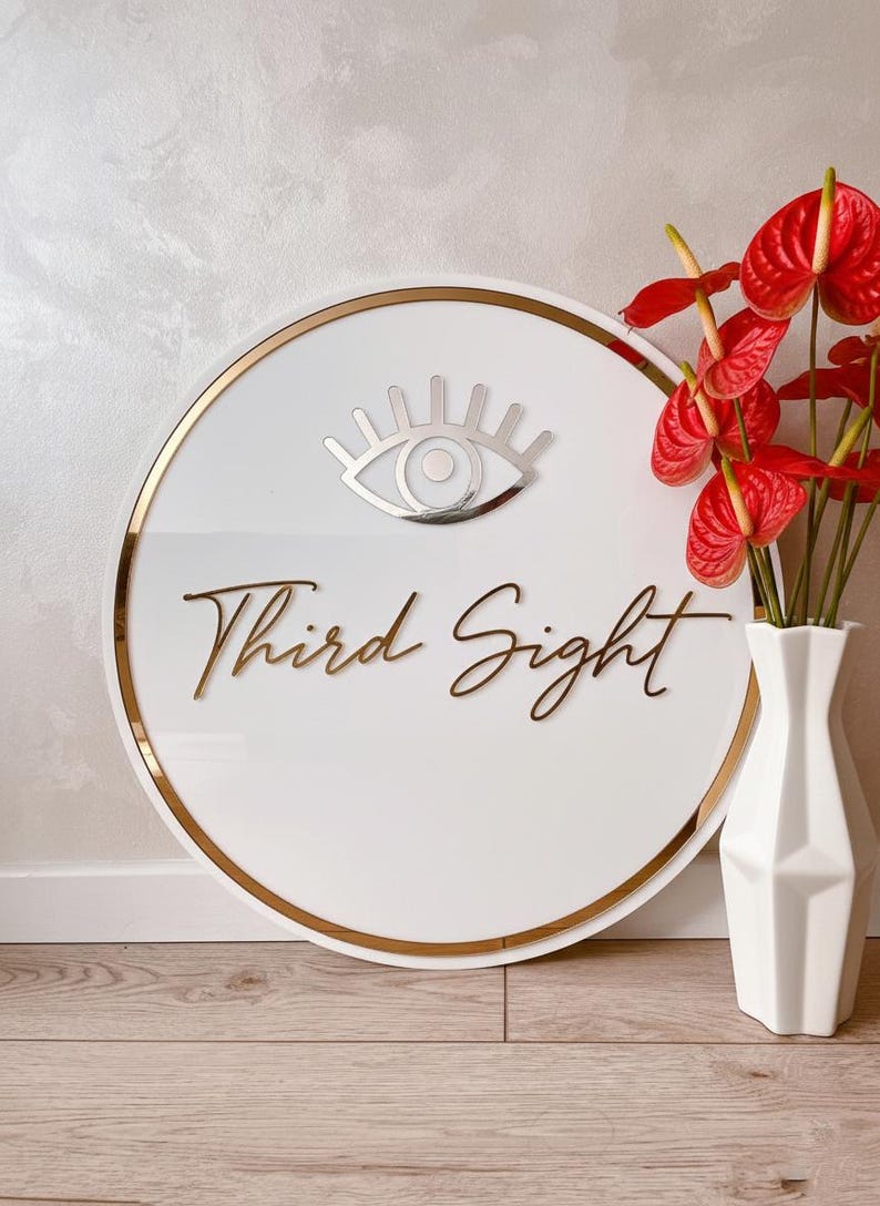 May include: A white circular sign with a gold border and the words "Third Sight" in gold script. A silver eye symbol is at the top. Red flowers in a white vase are to the right.