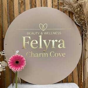 Pu&ograve; includere: Un cartello circolare beige con scritte dorate che recitano Beauty & Wellness Felyra Charm Cove e un'icona a forma di cuore sopra il testo. Una gerbera rosa brillante in un vaso di vetro trasparente si trova a sinistra del cartello.