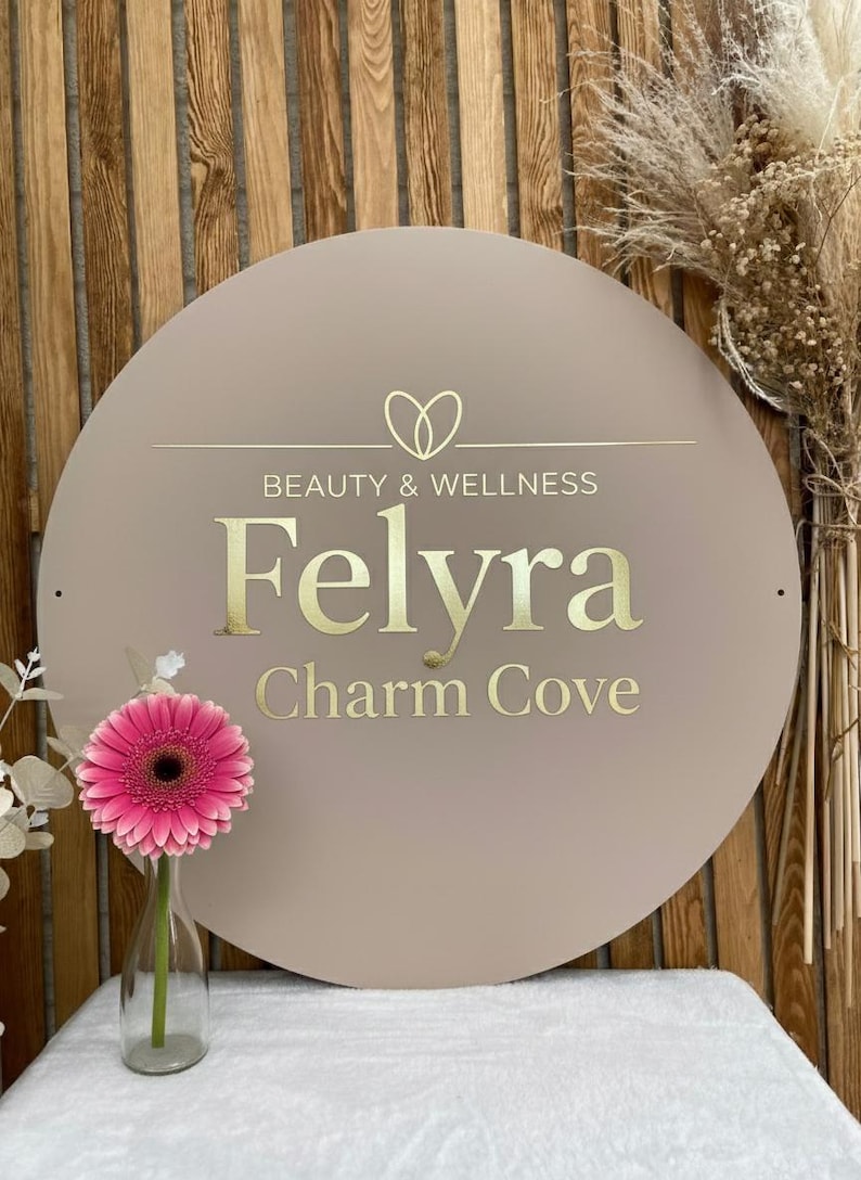 Pu&ograve; includere: Un cartello circolare beige con scritte dorate che recitano Beauty & Wellness Felyra Charm Cove e un'icona a forma di cuore sopra il testo. Una gerbera rosa brillante in un vaso di vetro trasparente si trova a sinistra del cartello.