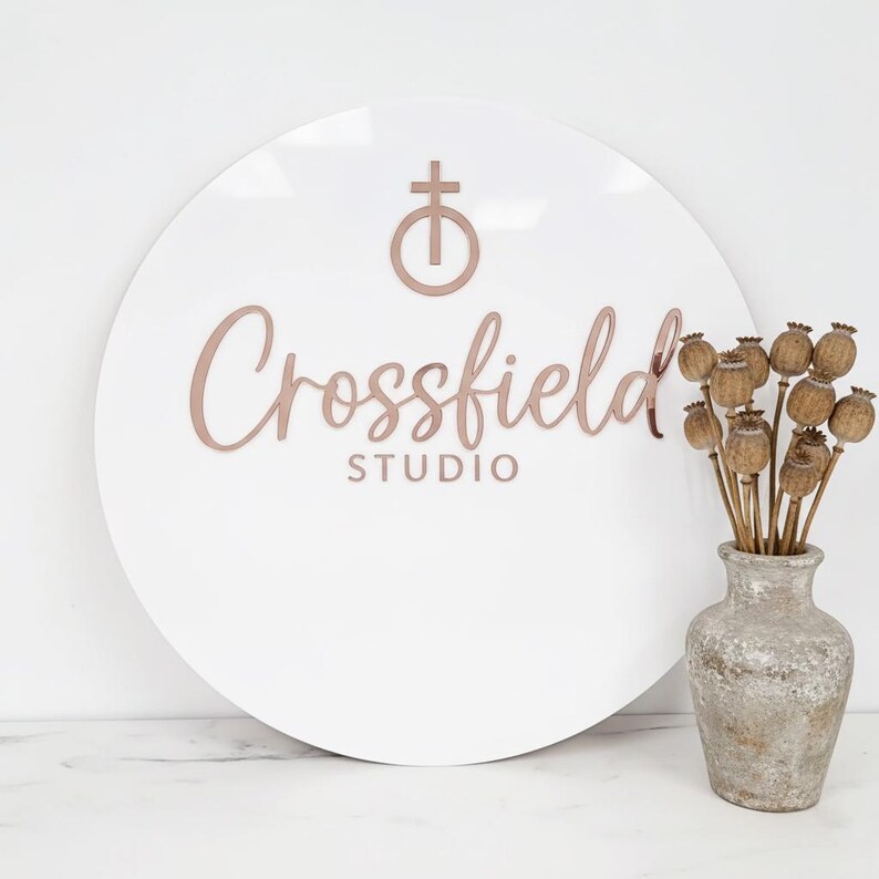 May include: A white, round sign with the text "Crossfield STUDIO" in a rose gold color. A rose gold cross symbol is above the text. A vase with dried seed pods sits to the right of the sign.
