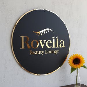 Pu&ograve; includere: Un'insegna circolare nera con bordo dorato a specchio e scritte dorate che recitano Rovella Beauty Lounge, caratterizzata da un grafico a ciglia dorato stilizzato sopra il testo. L'insegna &egrave; montata su una parete grigio chiaro testurizzata.