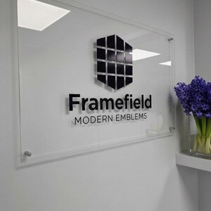 May include: Clear acrylic sign with the text "Framefield MODERN EMBLEMS" in black. The sign features a geometric black and silver emblem. The sign is mounted on a white wall with a small shelf holding a vase of purple flowers.