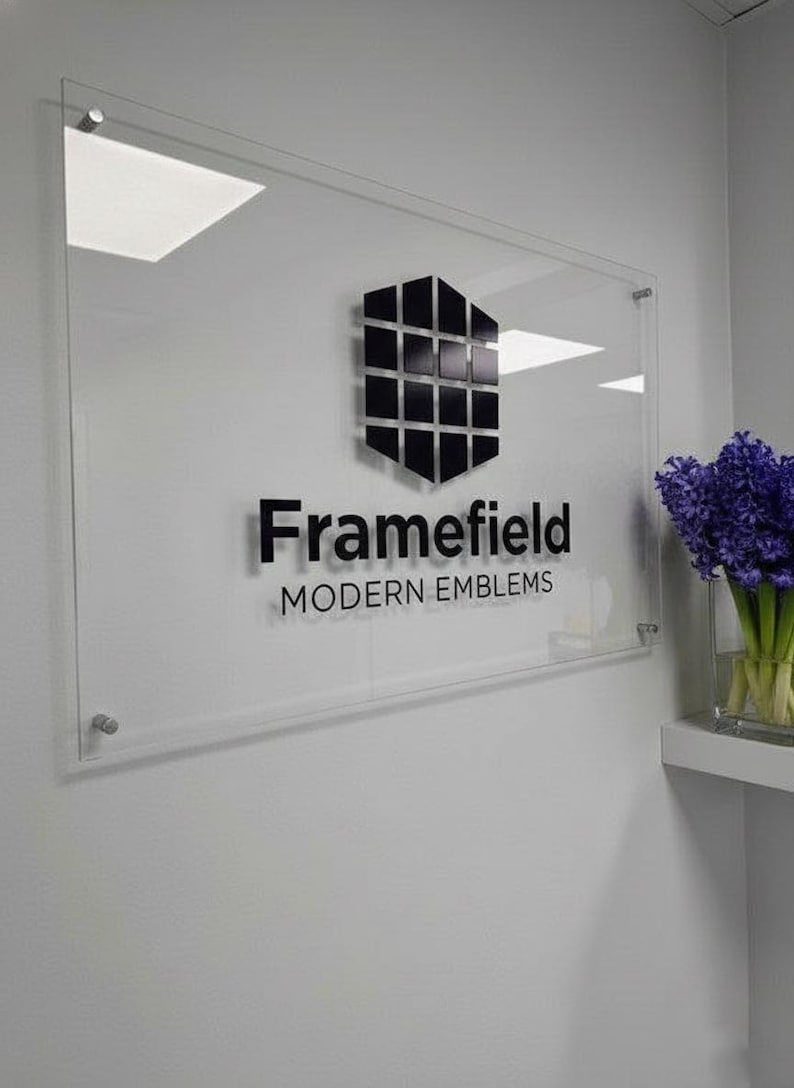 May include: Clear acrylic sign with the text "Framefield MODERN EMBLEMS" in black. The sign features a geometric black and silver emblem. The sign is mounted on a white wall with a small shelf holding a vase of purple flowers.