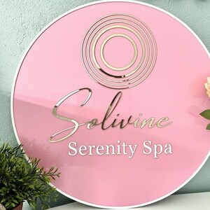 Pu&ograve; includere: Un cartello circolare rosa con bordo bianco che presenta un logo a cerchi concentrici in oro metallizzato nella parte superiore. Il testo Solivine &egrave; scritto in un carattere corsivo dorato, con le parole Serenity Spa in un carattere bianco.