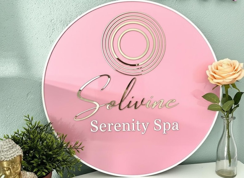 Pu&ograve; includere: Un cartello circolare rosa con bordo bianco che presenta un logo a cerchi concentrici in oro metallizzato nella parte superiore. Il testo Solivine &egrave; scritto in un carattere corsivo dorato, con le parole Serenity Spa in un carattere bianco.