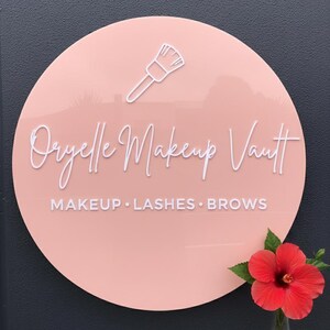 Pu&ograve; includere: Un'insegna circolare in acrilico rosa chiaro con il contorno bianco di un pennello da trucco, il testo "Oryelle Makeup Vault" in corsivo bianco e "MAKEUP &bull; LASHES &bull; BROWS" in un carattere sans-serif bianco.