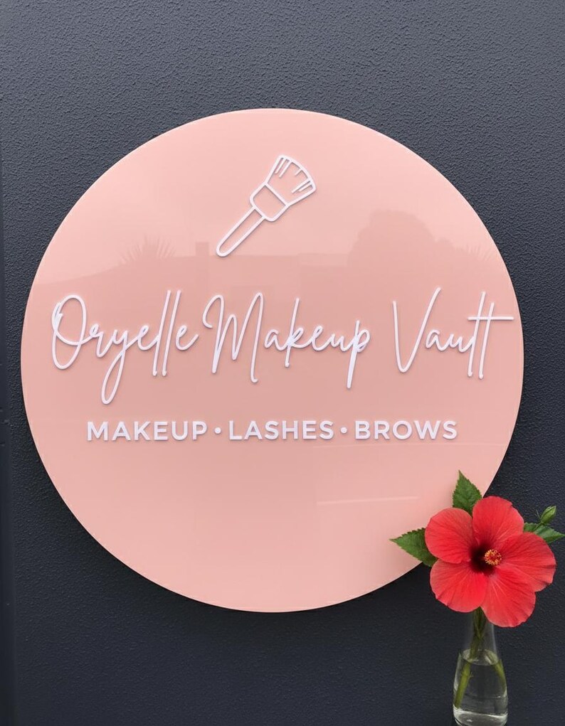 Pu&ograve; includere: Un'insegna circolare in acrilico rosa chiaro con il contorno bianco di un pennello da trucco, il testo "Oryelle Makeup Vault" in corsivo bianco e "MAKEUP &bull; LASHES &bull; BROWS" in un carattere sans-serif bianco.