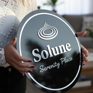 Może przedstawiać: Czarny owalny znak z białą obw&oacute;dką i srebrnymi literami. Znak głosi "Solune Serenity Place" i ma stylizowany wz&oacute;r na g&oacute;rze. Znak jest trzymany przez osobę.