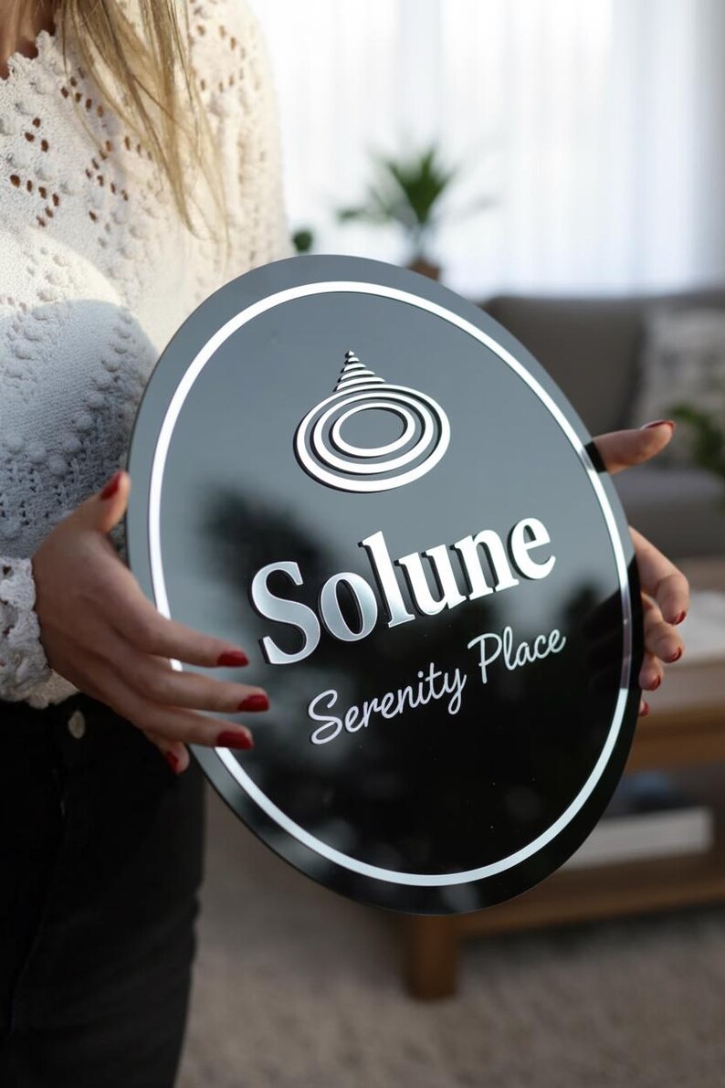 Może przedstawiać: Czarny owalny znak z białą obw&oacute;dką i srebrnymi literami. Znak głosi "Solune Serenity Place" i ma stylizowany wz&oacute;r na g&oacute;rze. Znak jest trzymany przez osobę.