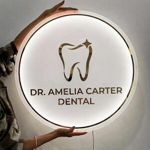 May include: A circular, illuminated sign featuring a gold-colored tooth icon with a small star at the top. The text "DR. AMELIA CARTER DENTAL" is printed in gold lettering below the tooth icon. The sign has a white, glowing border.