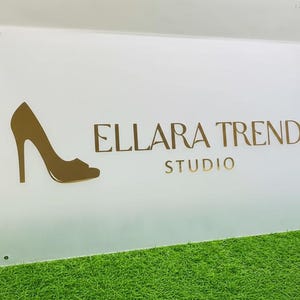 May include: A white rectangular sign with the text "ELLARA TREND STUDIO" in gold lettering. A gold high-heeled shoe silhouette is on the left. The sign is on a green surface.