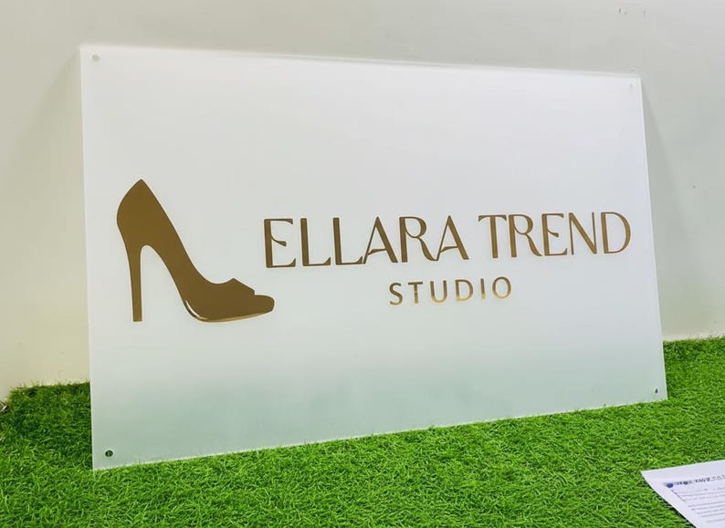 May include: A white rectangular sign with the text "ELLARA TREND STUDIO" in gold lettering. A gold high-heeled shoe silhouette is on the left. The sign is on a green surface.