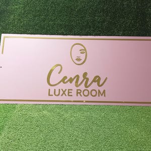 Może przedstawiać: Prostokątny, r&oacute;żowy znak ze złotymi obramowaniami i napisami. Znak zawiera nazwę "Cenra" w kursywie, z napisem "LUXE ROOM" poniżej. Minimalistyczna grafika twarzy znajduje się nad nazwą.