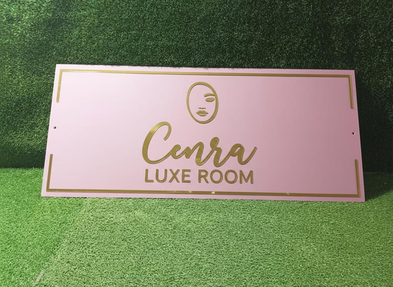 Może przedstawiać: Prostokątny, r&oacute;żowy znak ze złotymi obramowaniami i napisami. Znak zawiera nazwę "Cenra" w kursywie, z napisem "LUXE ROOM" poniżej. Minimalistyczna grafika twarzy znajduje się nad nazwą.