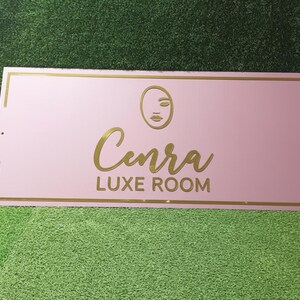 May include: A pink rectangular sign with gold detailing. The sign displays the name "Cenra" in a cursive font, with "LUXE ROOM" underneath. A minimalist face graphic is above the name.