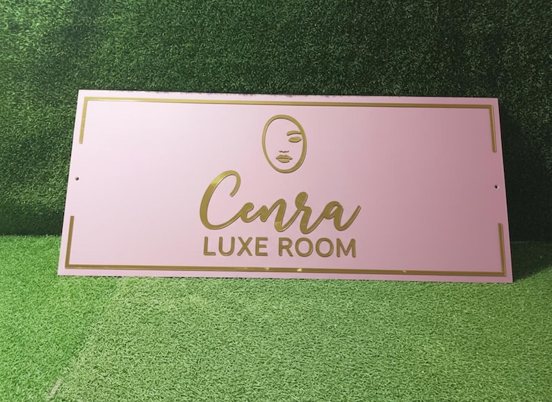 May include: A pink rectangular sign with gold detailing. The sign displays the name "Cenra" in a cursive font, with "LUXE ROOM" underneath. A minimalist face graphic is above the name.
