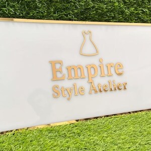 Może przedstawiać: Prostokątny znak z białym tłem i złotą obw&oacute;dką. Znak zawiera słowa "Empire Style Atelier" w złotych literach, wraz ze złotą sylwetką sukienki. Znak jest umieszczony na tle zielonej trawy.