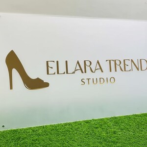 Może przedstawiać: Biały prostokątny znak z napisem "ELLARA TREND STUDIO" w złotych literach. Z lewej strony znajduje się złota sylwetka buta na wysokim obcasie. Znak znajduje się na zielonej powierzchni.