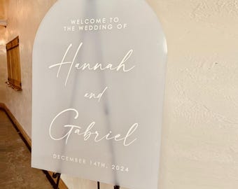 May include: An arched, frosted acrylic wedding sign featuring white text that reads: WELCOME TO THE WEDDING OF Hannah and Gabriel DECEMBER 14TH, 2024. The sign is mounted on a thin black metal easel stand, displaying a smooth, semi-transparent surface.