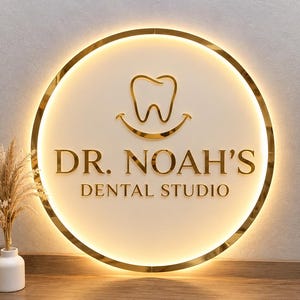 May include: A circular wall sign with a gold metallic frame and backlighting. The sign features a gold tooth icon with a smile underneath, and the text "DR. NOAH'S DENTAL STUDIO" in gold lettering against a smooth, off-white circular background.