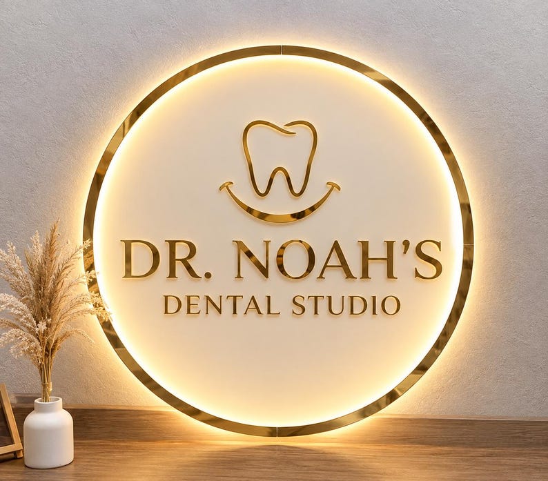 May include: A circular wall sign with a gold metallic frame and backlighting. The sign features a gold tooth icon with a smile underneath, and the text "DR. NOAH'S DENTAL STUDIO" in gold lettering against a smooth, off-white circular background.