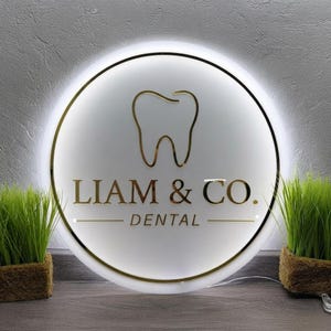 May include: A circular, illuminated wall sign featuring a gold-colored tooth icon centered above the text LIAM & CO. DENTAL in gold lettering. The sign has a white face with a thin gold border and emits a soft white glow from behind its edges.