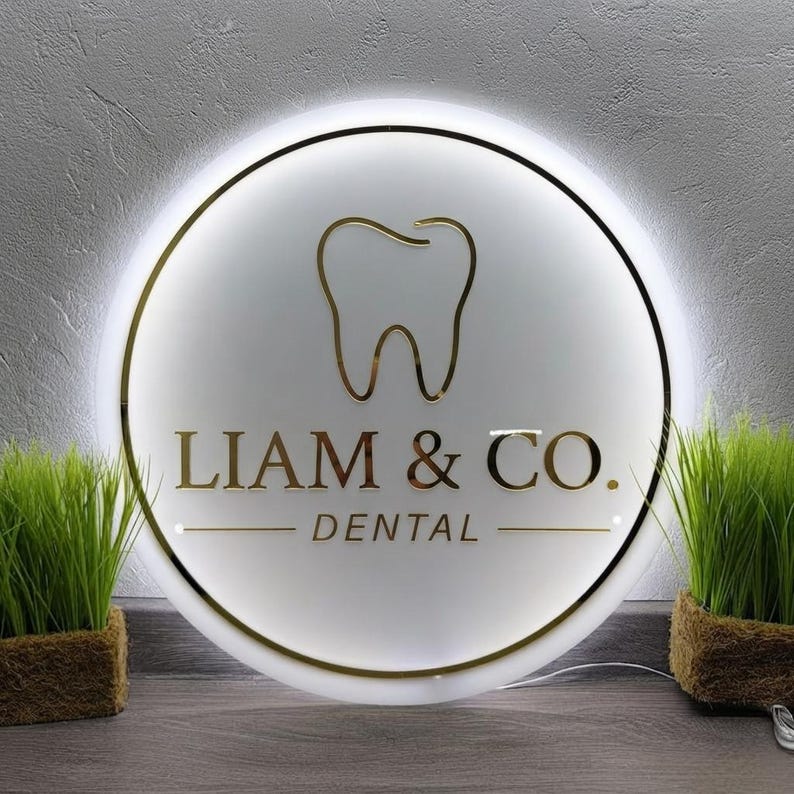 May include: A circular, illuminated wall sign featuring a gold-colored tooth icon centered above the text LIAM & CO. DENTAL in gold lettering. The sign has a white face with a thin gold border and emits a soft white glow from behind its edges.