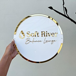 May include: A round white sign with gold trim and lettering. The sign reads "Soft River Balance Lounge" in gold lettering. A gold teardrop shape is above the words "Soft River". The sign is held by a hand.