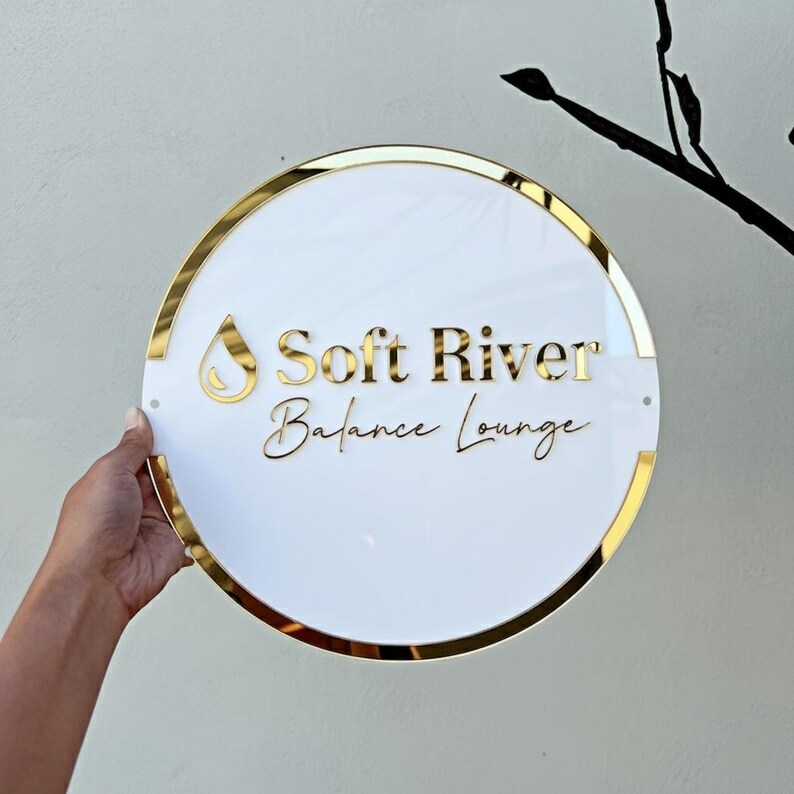 May include: A round white sign with gold trim and lettering. The sign reads "Soft River Balance Lounge" in gold lettering. A gold teardrop shape is above the words "Soft River". The sign is held by a hand.