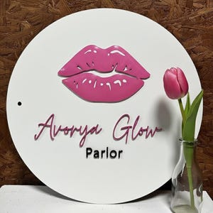 Pu&ograve; includere: Un cartello circolare bianco con una grafica in rilievo di labbra rosa nella parte superiore. Sotto le labbra, il testo recita Avorya Glow in un carattere corsivo rosa, con la parola Parlor in un carattere nero pi&ugrave; piccolo. Un foro &egrave; visibile.